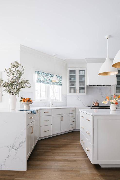 A white angled kitchen boasts a white island donning brass pulls and a marble top lit by two Aerin Agnes pendants. A marble waterfall top accents a peninsula.