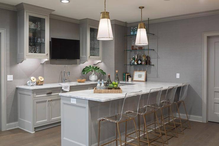Gray basement bar boasts Gabby King Barstools placed at a gray peninsula topped with a white and gray countertop lit by two glass and brass pendants. The stools face a television mounted to a gray grasscloth wallpapered wall between glass front cabinets finished with glass shelves. The television is positioned above a sink with a stainless steel pull out faucet fitted above gray cabinets. The bar is completed with glass and bras wall shelves showcasing an assortment of spirits.