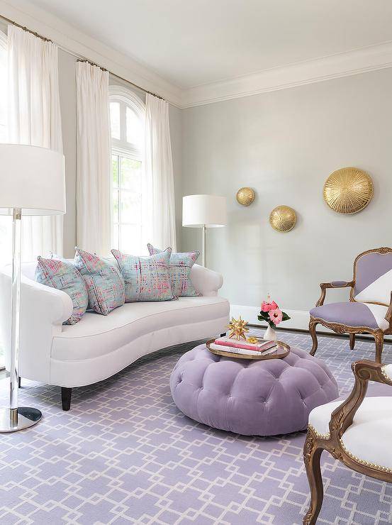 Arched windows framed by a light gray wall and covered in white curtains are positioned behind a white curved sofa topped with pink and blue pillows and placed on a purple chain link rug between glass and nickel floor lamps. White and purple French chairs sit facing a purple velvet tufted ottoman coffee table placed in front of the sofa.