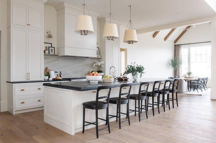 Dalston Hanging Shades hang over a long white kitchen island contrasted with a honed black marble countertop finished with a sink and complemented with black vintage style stools.