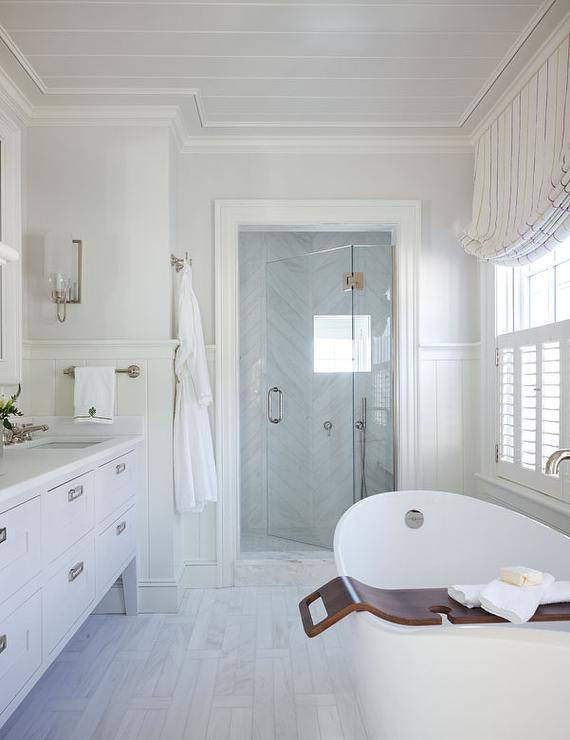 Narrow marble tile floors, white herringbone shower wall tiles, a white free-standing sink with polished nickel drawer pulls lit by a polished nickel wall sconce, a walk-in shower boasting a frameless glass shower door and an oval bathtub with a teak tray under a window with gray striped roman shades are featured in this stunning long master bathroom.